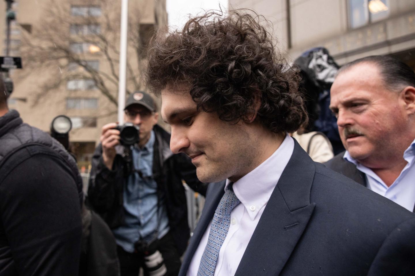 FTX founder Sam Bankman-Fried looks down as he exits the courthouse as media throng around him.