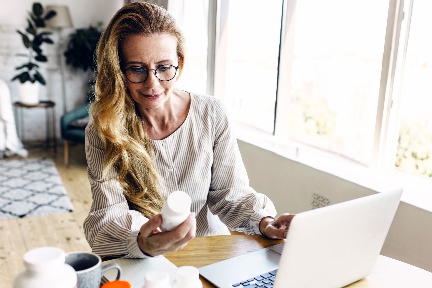 woman sitting in living-room at table using laptop, holding bottle of medication