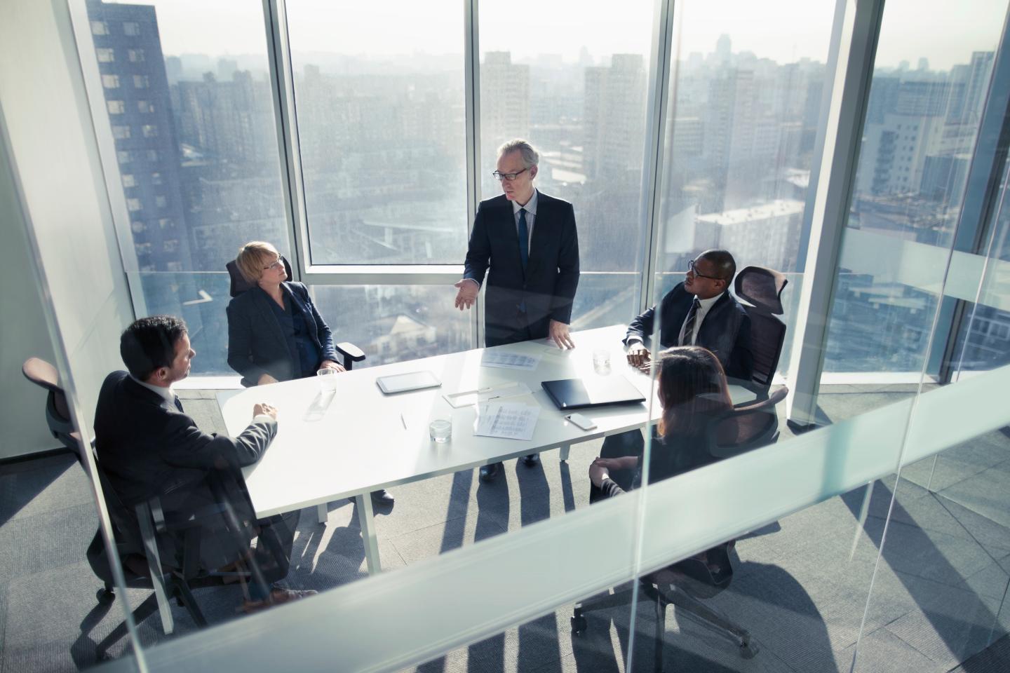Mature senior adult businessman leader boss CEO executive with group of business people sitting together in conference meeting room in contemporary modern office bright sunny daylight talking discussing planning organizing strategy well dressed suit and tie teamwork cooperation diversity multi-ethnic problems solutions unity cityscape downtown urban