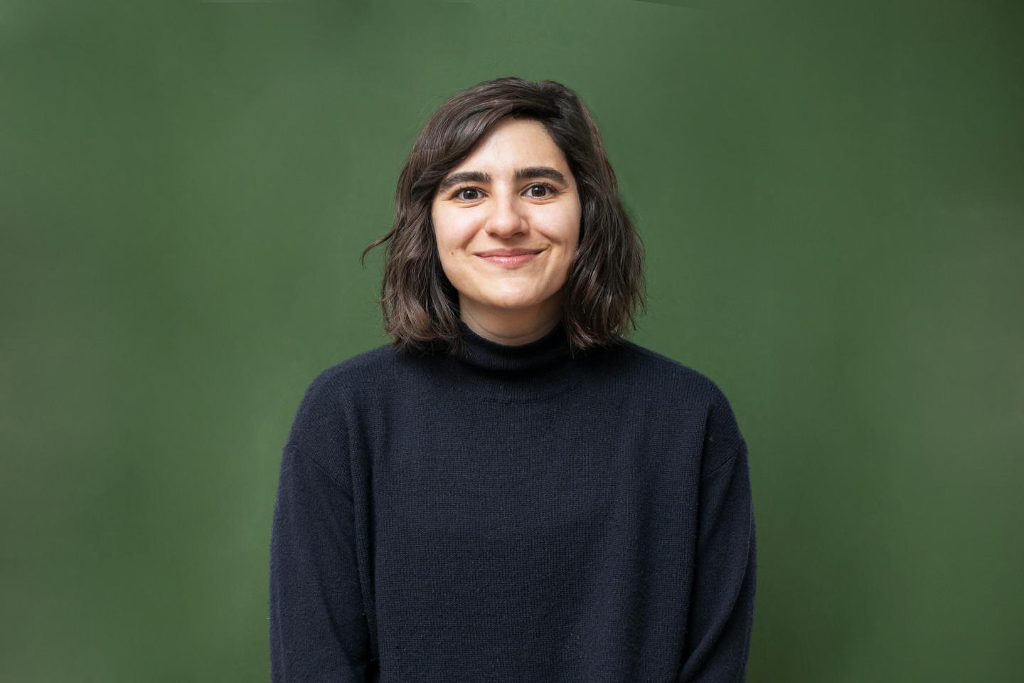 A young woman wearing a black sweater smiles as she poses in front of a dark green background