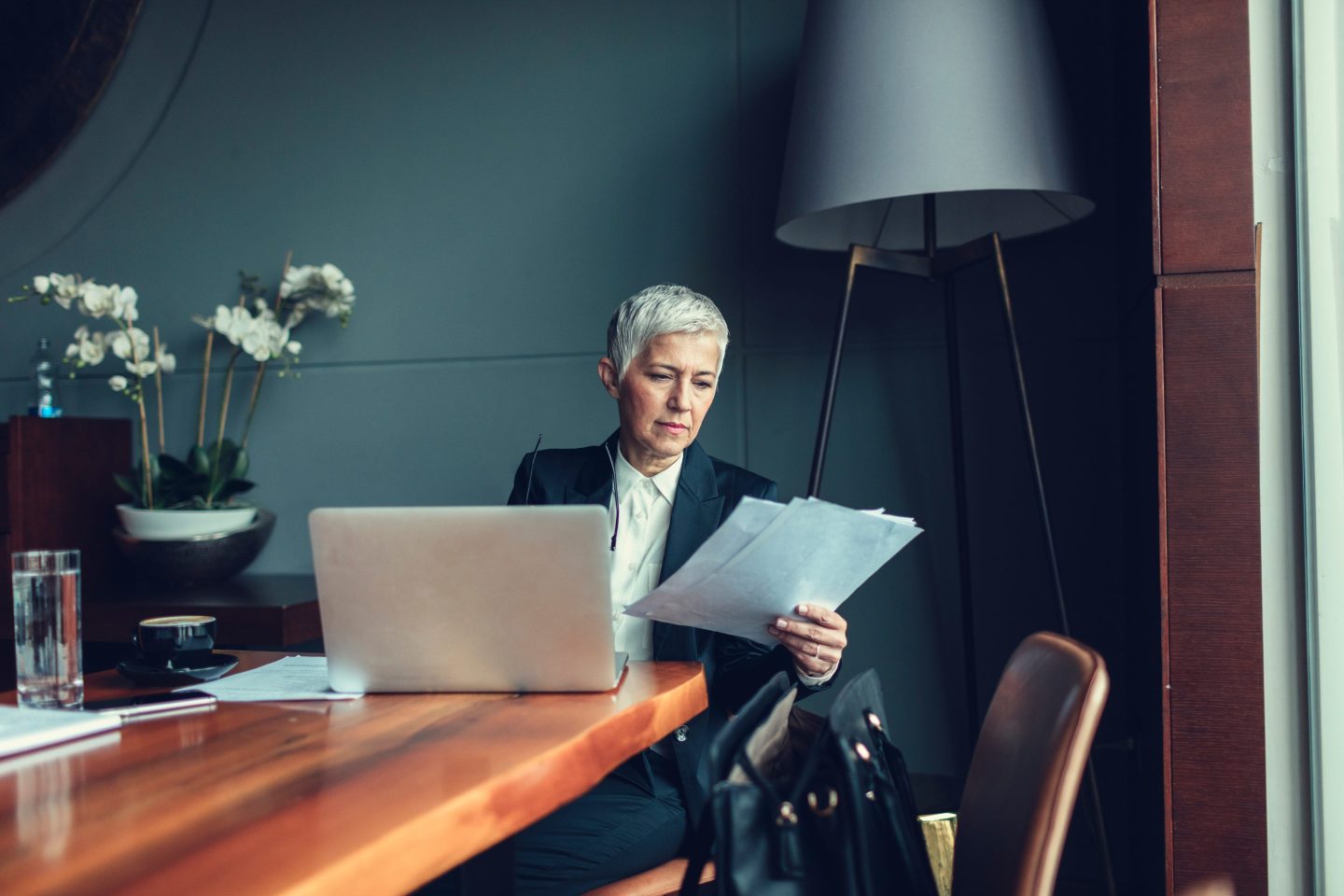 Mature businesswoman working, sitting in restaurant or office. In front of her, on the table, is laptop. She is holding papers and looks worried.