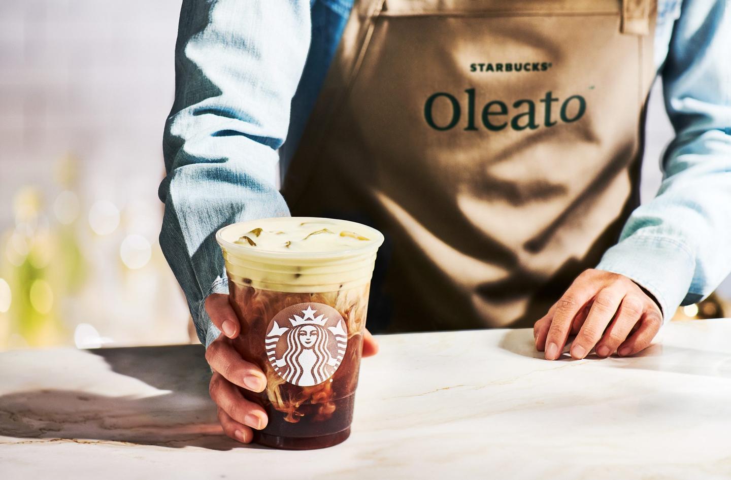 Image of a Starbucks barista wearing a long-sleeved blue shirt and a tan apron holding a Starbucks drink