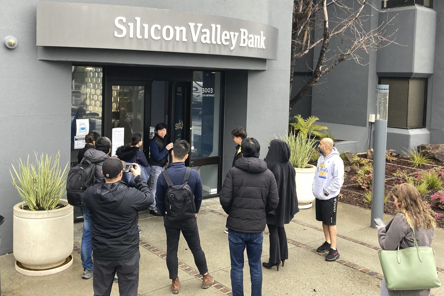 A person from inside Silicon Valley Bank, middle rear, talks to people waiting outside of an entrance to Silicon Valley Bank in Santa Clara, Calif., on March 10, 2023.
