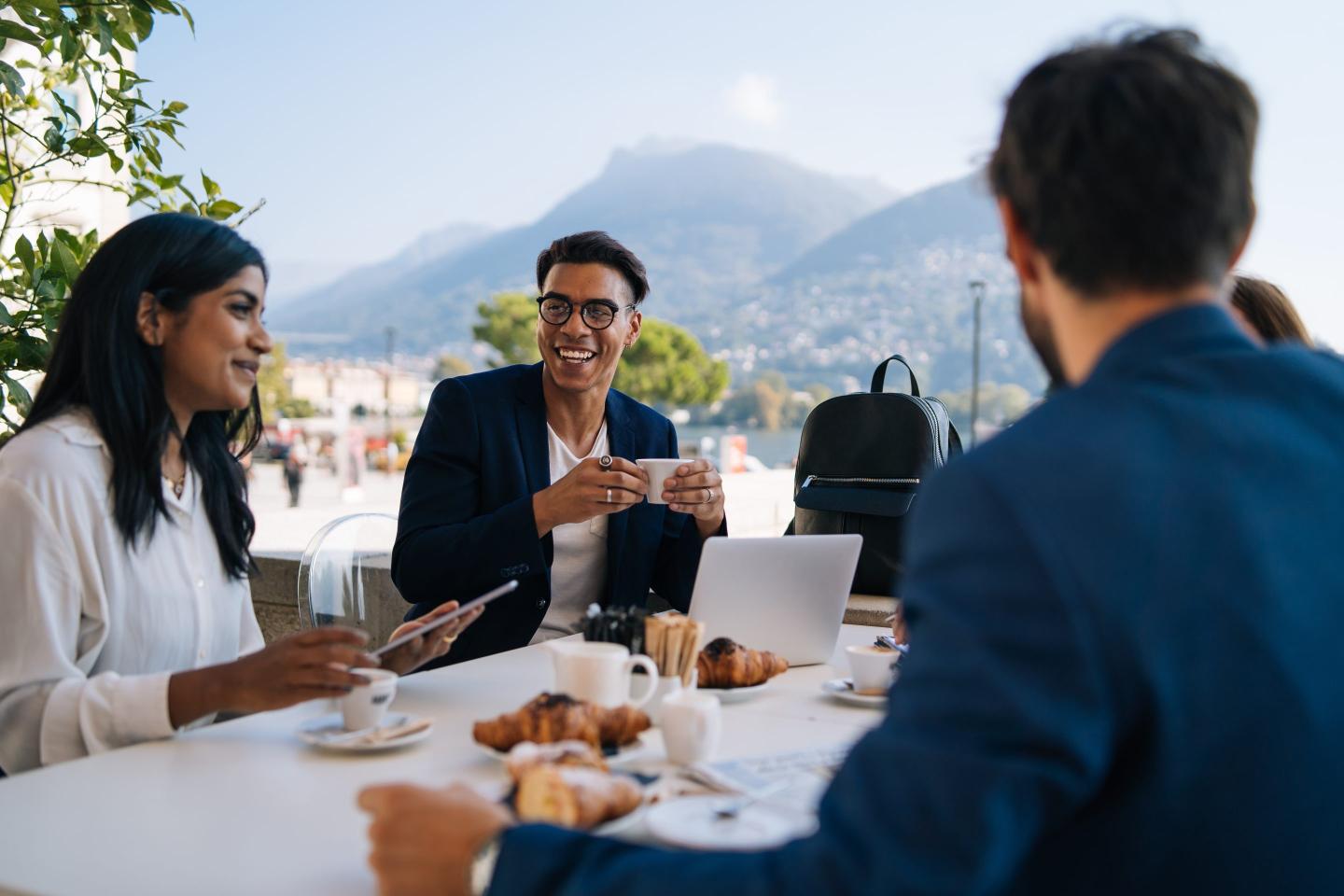Four people dressed in business casual attire sit at a table outside, eating and drinking food while working on their phones and laptops.