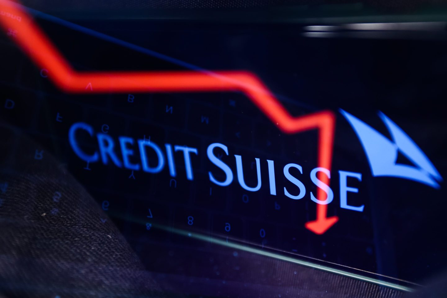 An illustrative stock chart displayed on a laptop screen and Credit Suisse logo displayed on a phone screen.