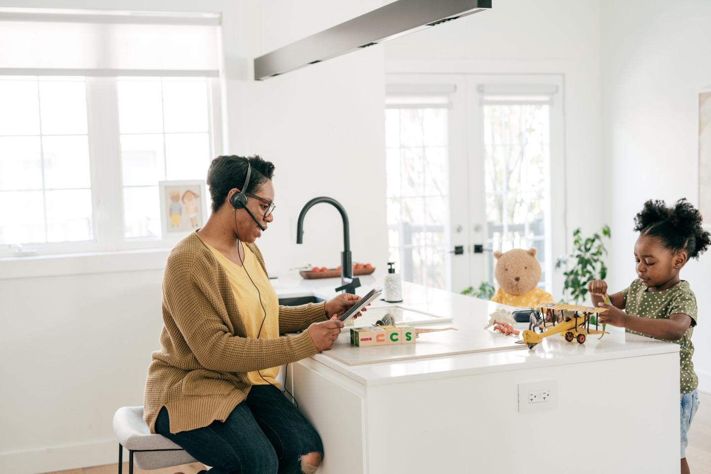 A woman wearing a headset sits at a kitchen island taking work calls, while her daughter plays with toys on the other side.