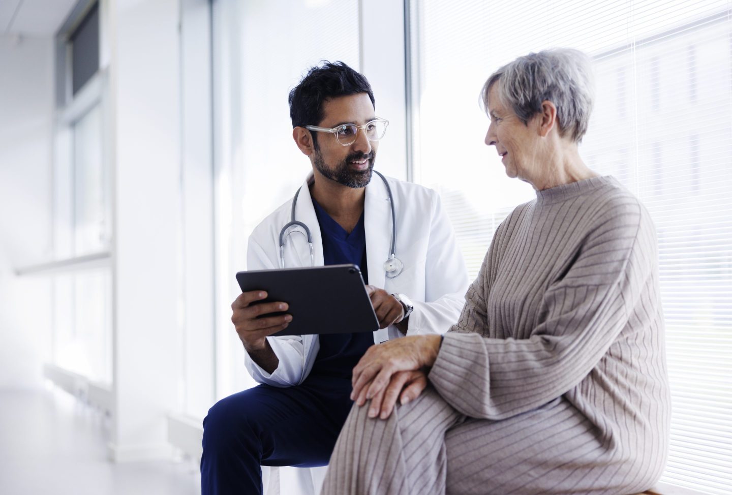 A male doctor holding a tablet and female patient sit on a bench in conversation.