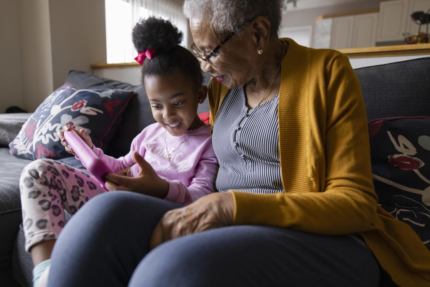 Grandmother sits with granddaughter on couch looking at digital tablet.