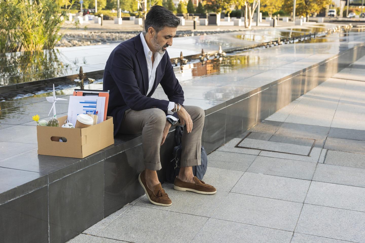 Sad businessman with office supplies in box sitting on wall