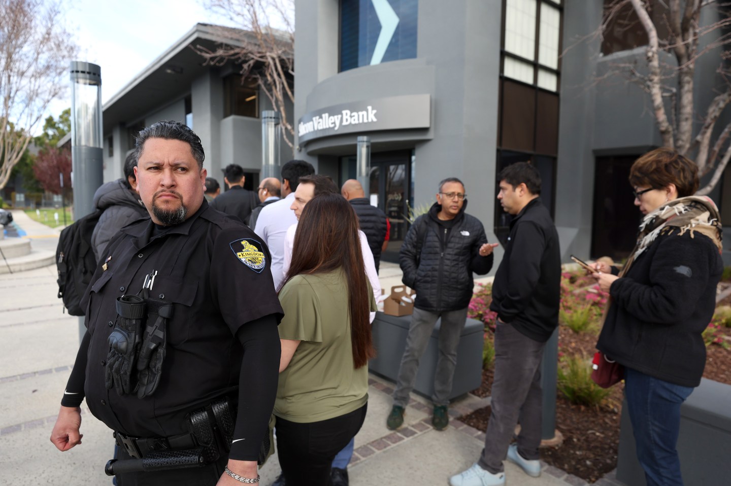 A security guard monitors a line of people outside of a Silicon Valley Bank office on March 13, 2023 in Santa Clara, California. Days after Silicon Valley Bank collapsed, customers are lining up to try and retrieve their funds from the failed bank. The Silicon Valley Bank failure is the second largest in U.S. history.