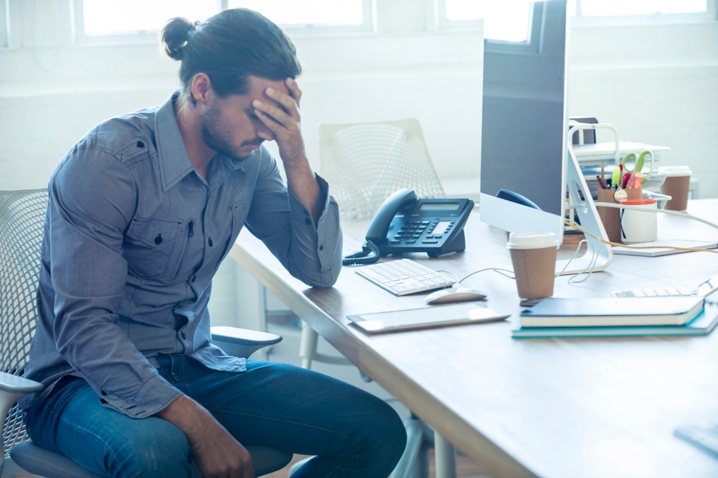 Stressed business man at the office. He is casually dressed and looking distraught. He looks very uncomfortable and could also have a headache. He is has his head in his hand and looks very upset. Hi is sitting at a desk with a computer and phone. Copy space.