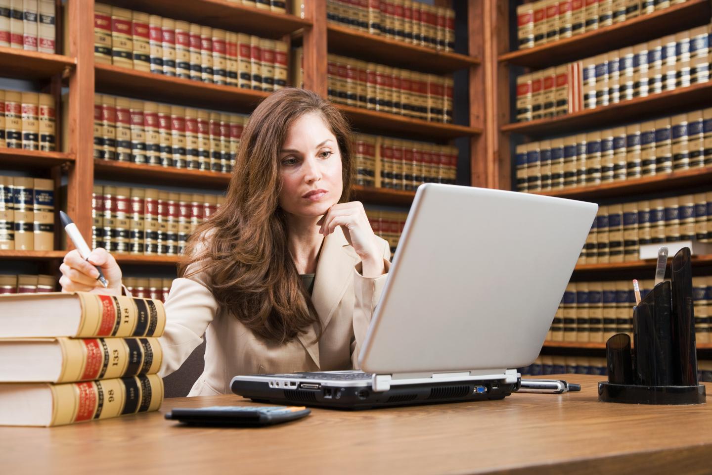 Photo of a woman sitting at a desk looking at a computer in front of a library shelf full of legal books.