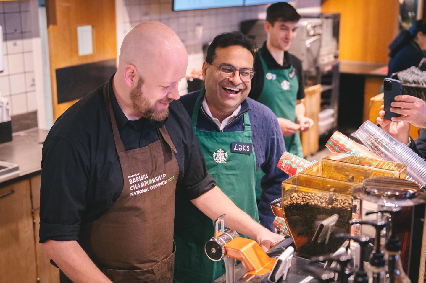 Starbucks CEO Laxman Narasimhan, wearing a green barista apron, works behind a Starbucks counter