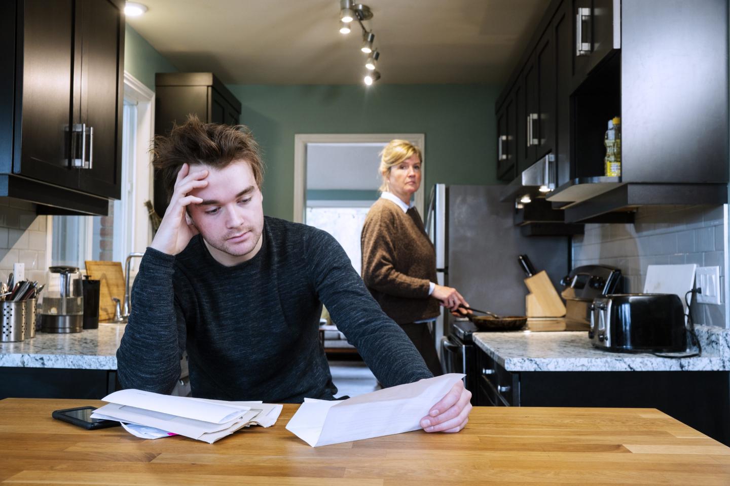 A young man sitting at home stressed by his mounting debts. His mother looks on