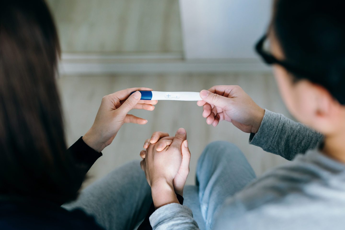 High angle view of an affectionate young Asian couple sitting on the bed, holding hands and holding a positive pregnancy test together.