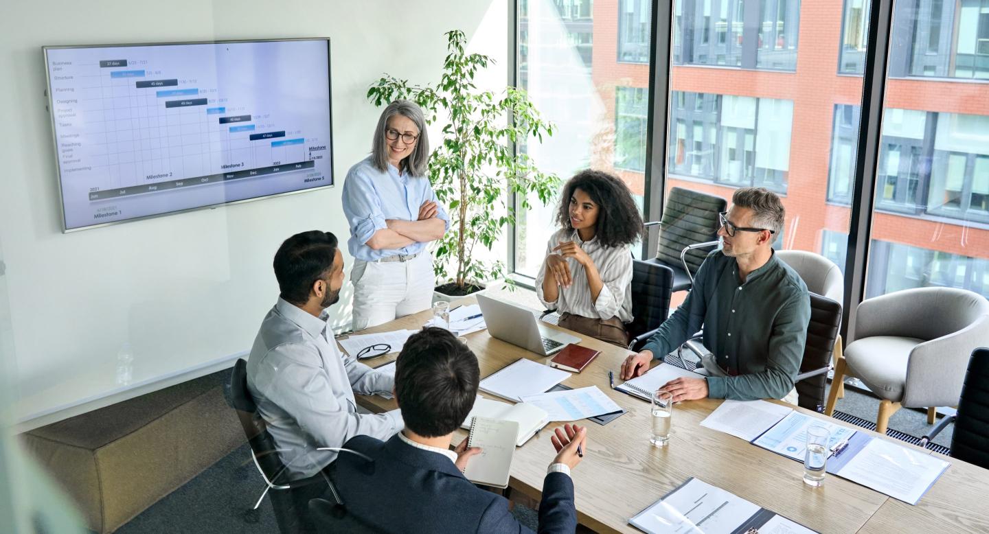 Senior female leader and multicultural business people discussing company presentation at boardroom table. Diverse corporate team working together in modern meeting room office. Top view through glass