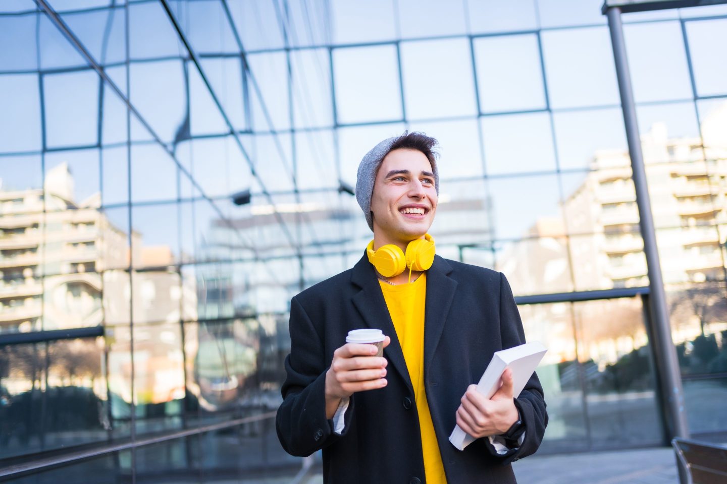 Young businessman in black jacket and yellow t-shirt heading to work with headphones. With agenda and a coffee take away outside the office