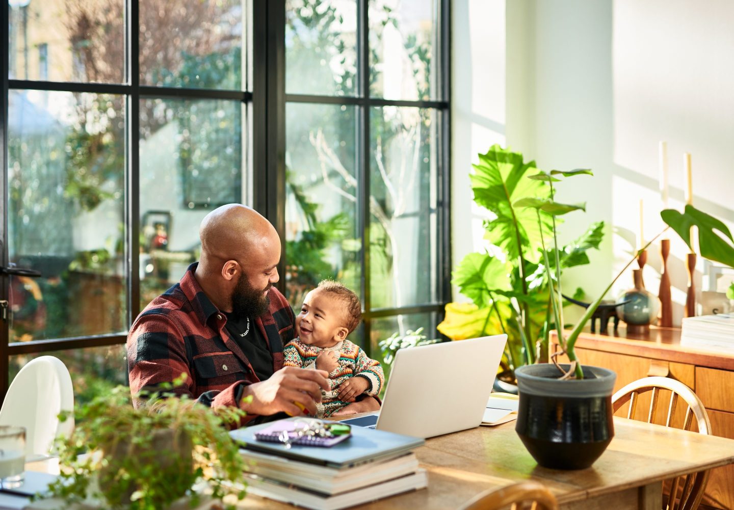 Black father smiling face to face with baby son in front of laptop in home office