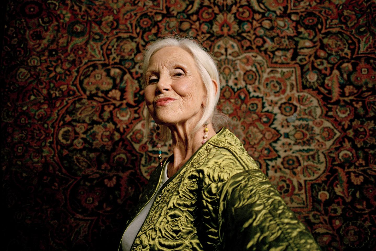 Side view of elderly woman wearing golden jacket puckering by wall hanging rug.
