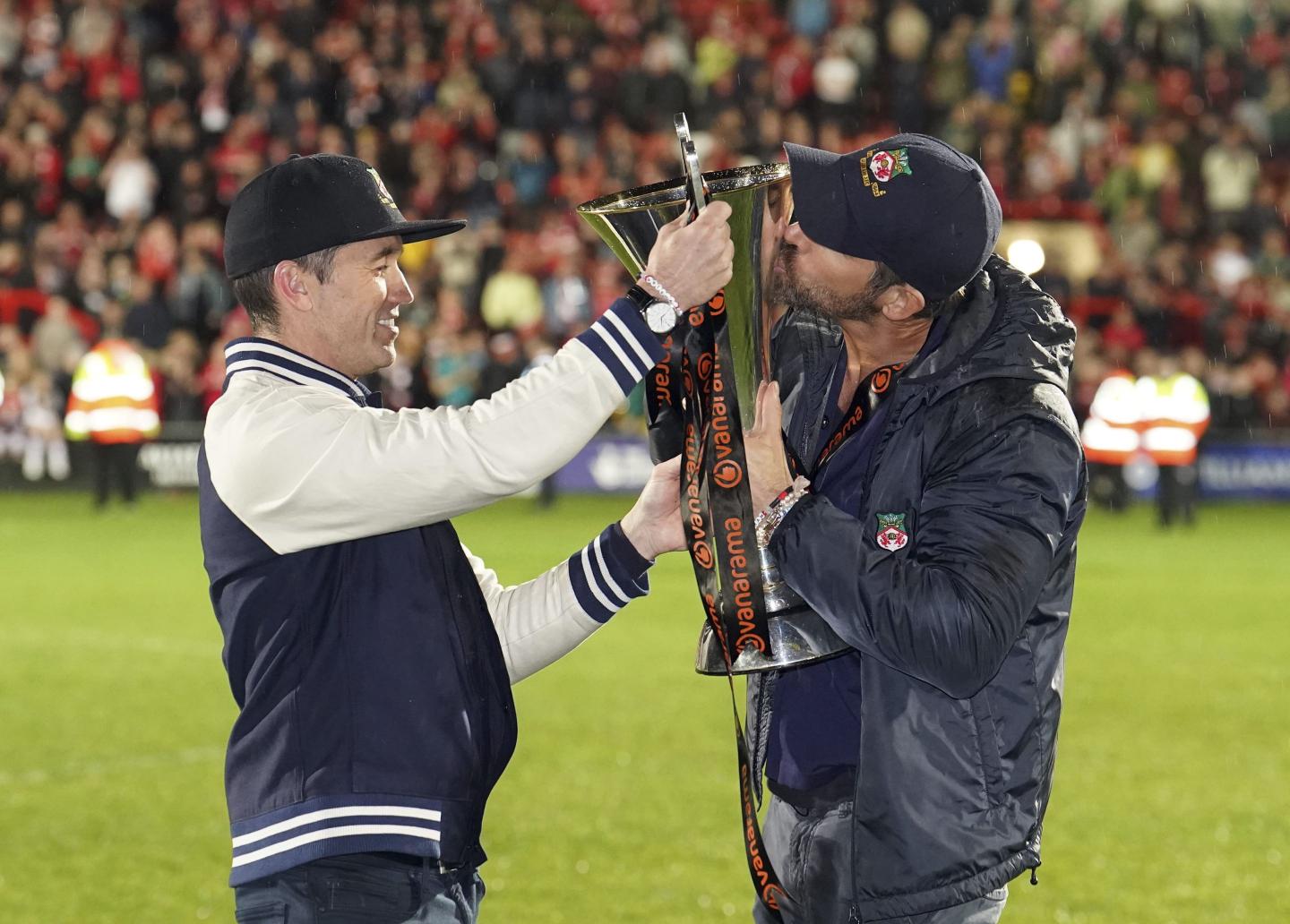 Wrexham co-owners Rob McElhenney, left, and Ryan Reynolds celebrate with the trophy after their team clinched promotion following the National League soccer match between Wrexham and Boreham Wood at The Racecourse Ground, in Wrexham, Wales, Saturday April 22, 2023. 