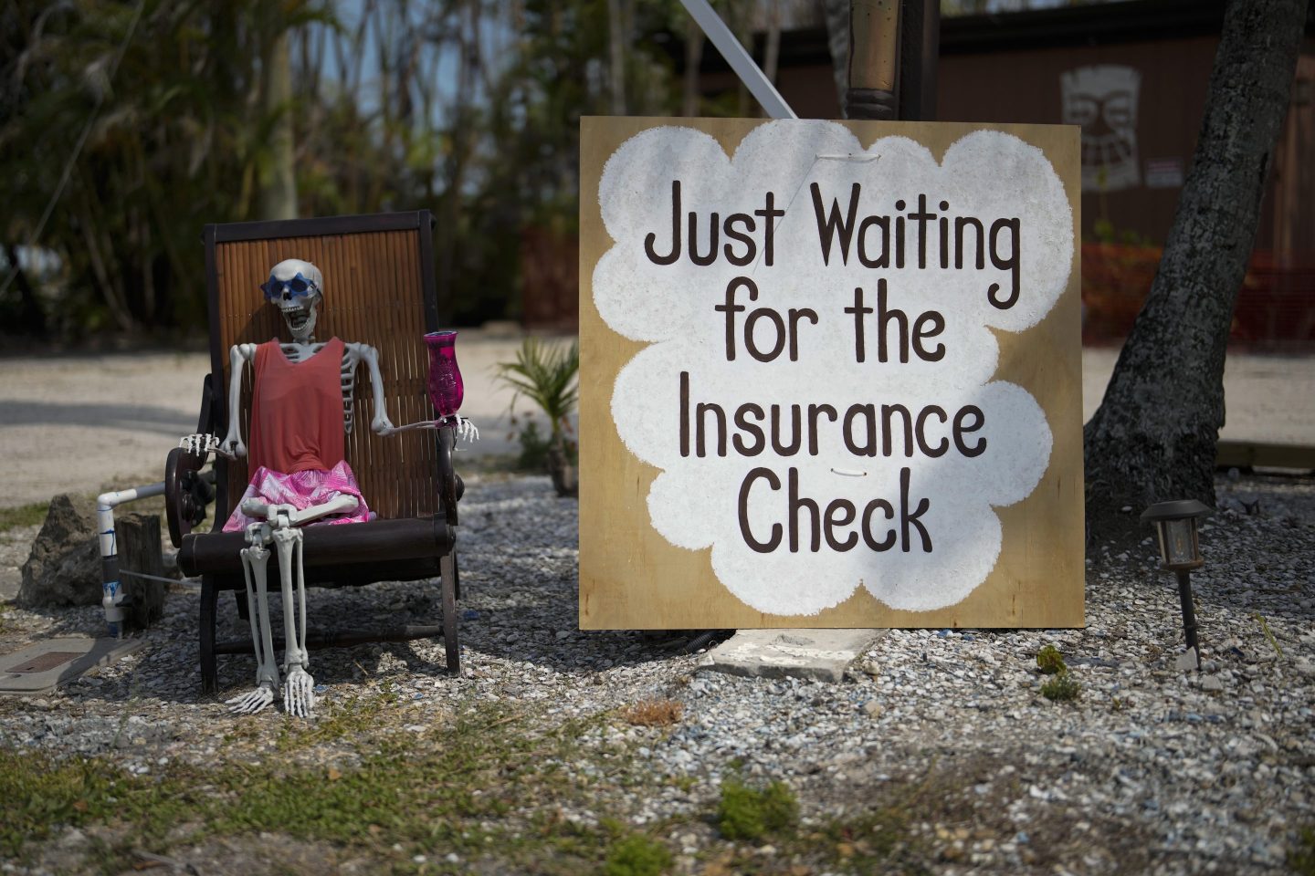 A skeleton in sunglasses sits beside a sign reading "Just waiting for the insurance check," outside the closed Kona Kai Motel on Sanibel Island, Fla., on May 11, 2023.
