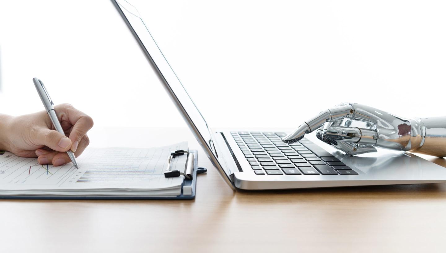 Human hand holding a pen to paper on the left; a robotic metal hand typing on a laptop to the right