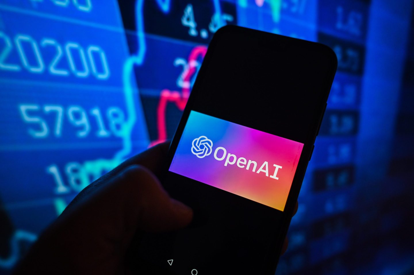 A phone screen with OpenAI's logo in front of a large monitor displaying financial data
