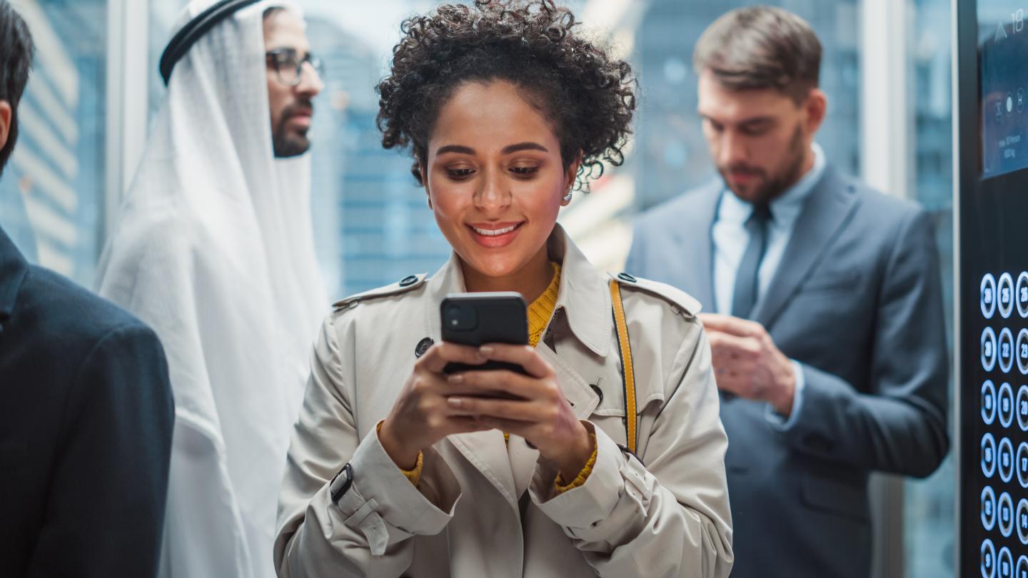 A woman stands in a crowded lift smiling at her phone.