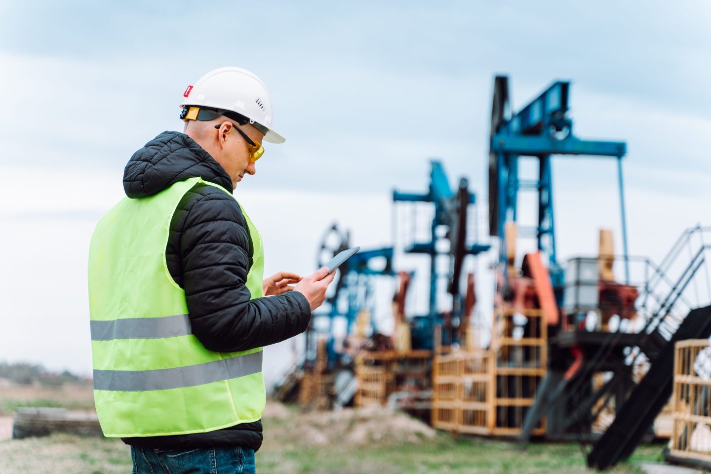 An man in a high visibility vest looks at a tablet while visiting an oil well.