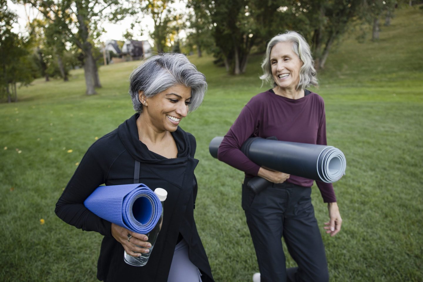 Two women carrying yoga mats walk in a park