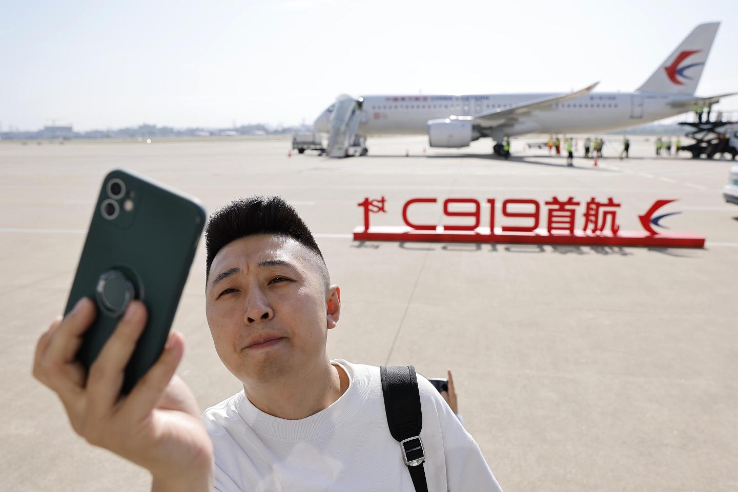 A man takes a selfie in front of a sign celebrating the first commercial flight of the C919, as the plane sits in the background.