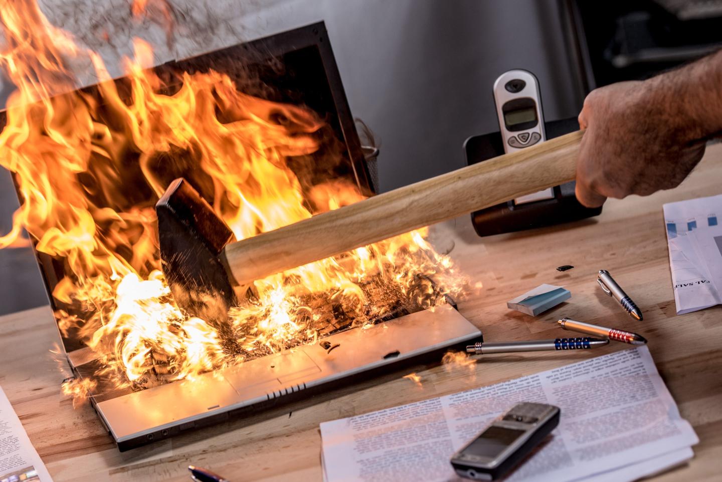 Businessman hitting hammer on burning laptop in office.