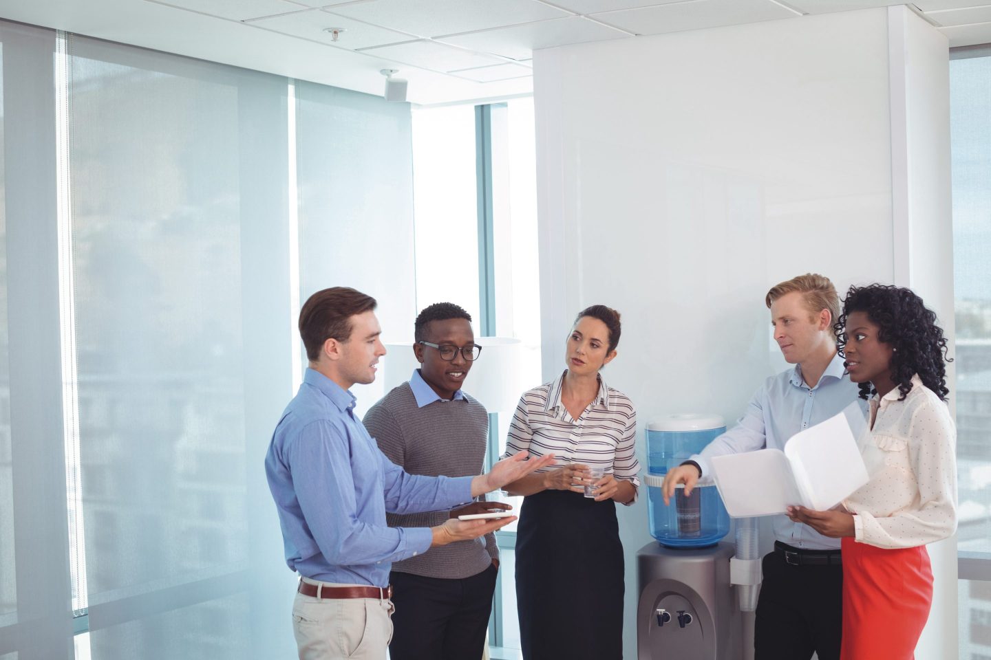 Five people dressed in business casual attire stand around a water cooler, holding a discussion.