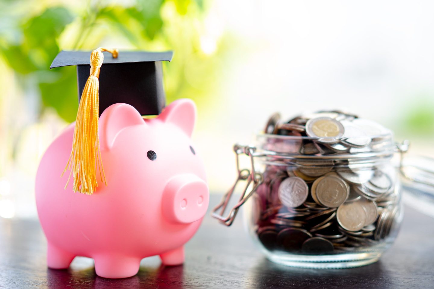 Piggy bank with a graduation cap is on a table next to a jar of coins.