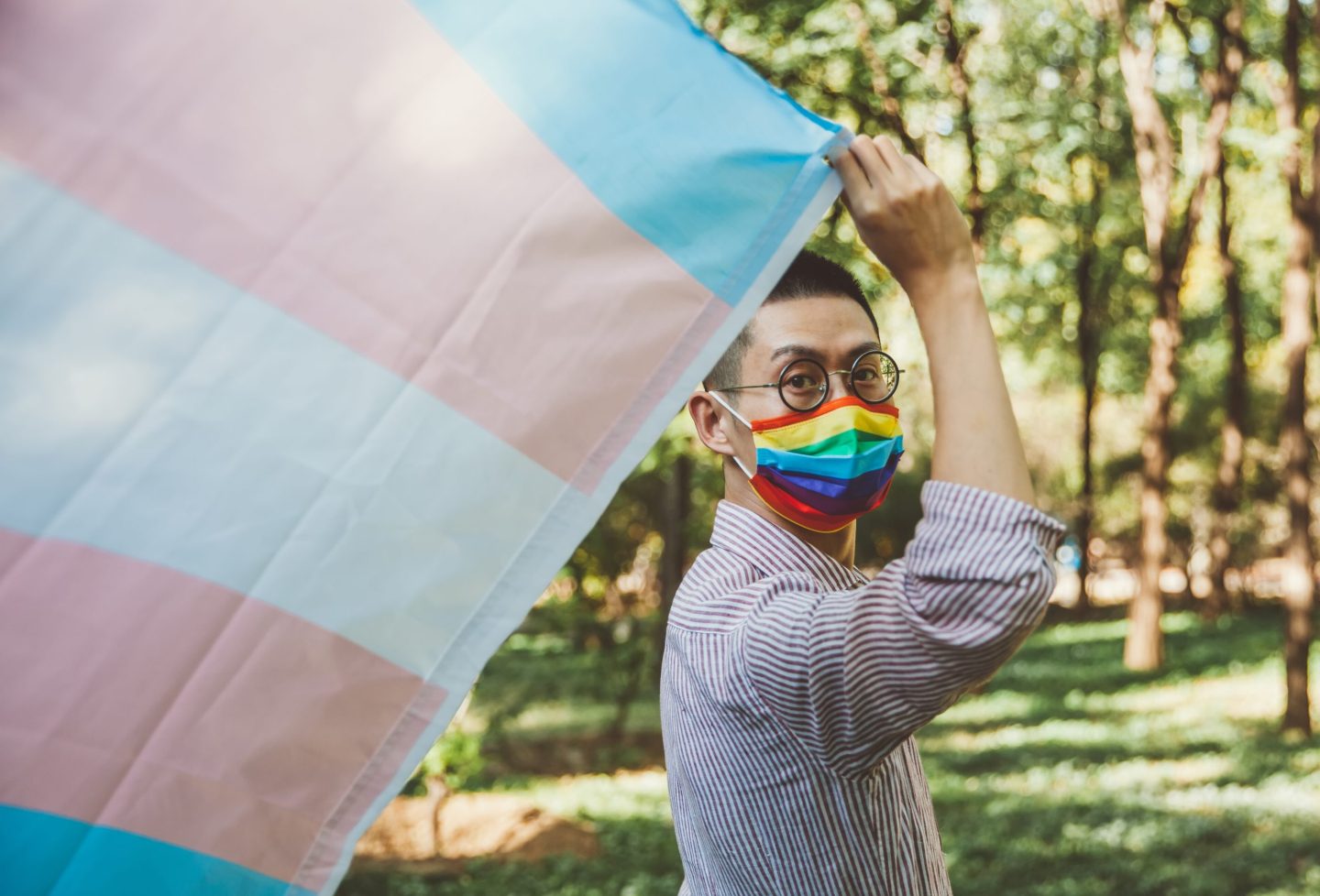 closeup of a perosn holding a transgender pride flag and wearing a rainbow mask