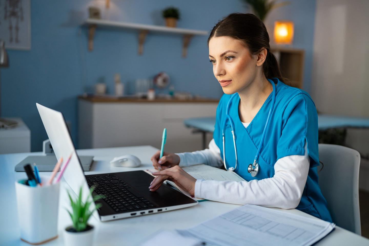 Woman in scrubs works on a laptop.