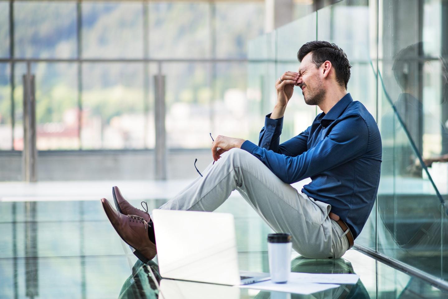 Frustrated young man with laptop sitting on the floor in the modern building, holding his head.