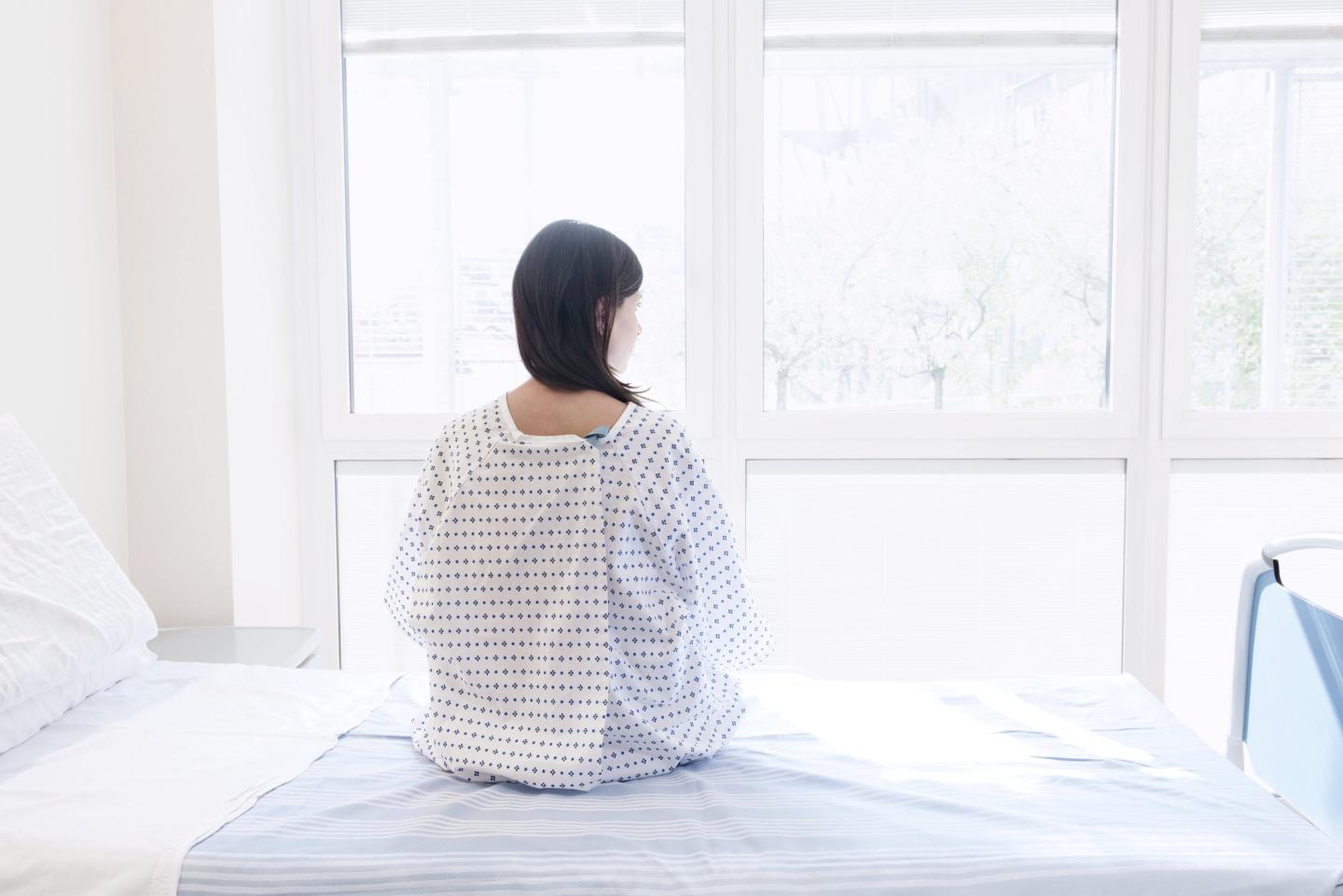 A woman sitting in a hospital gown on a bed, looking out the window