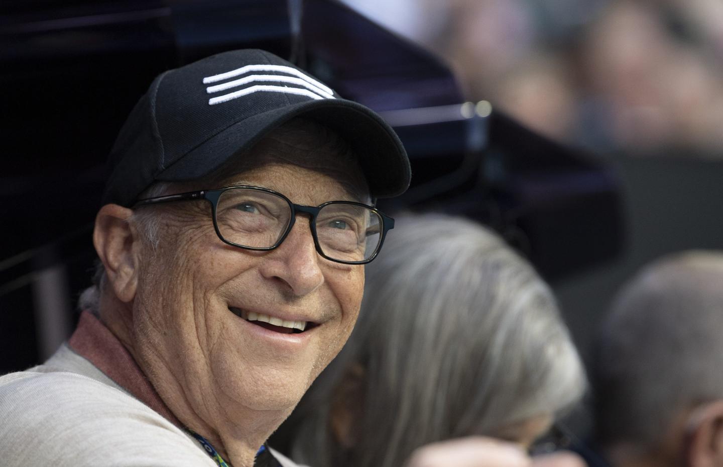 Bill Gates wears a cap and smiles while watching a tennis match.