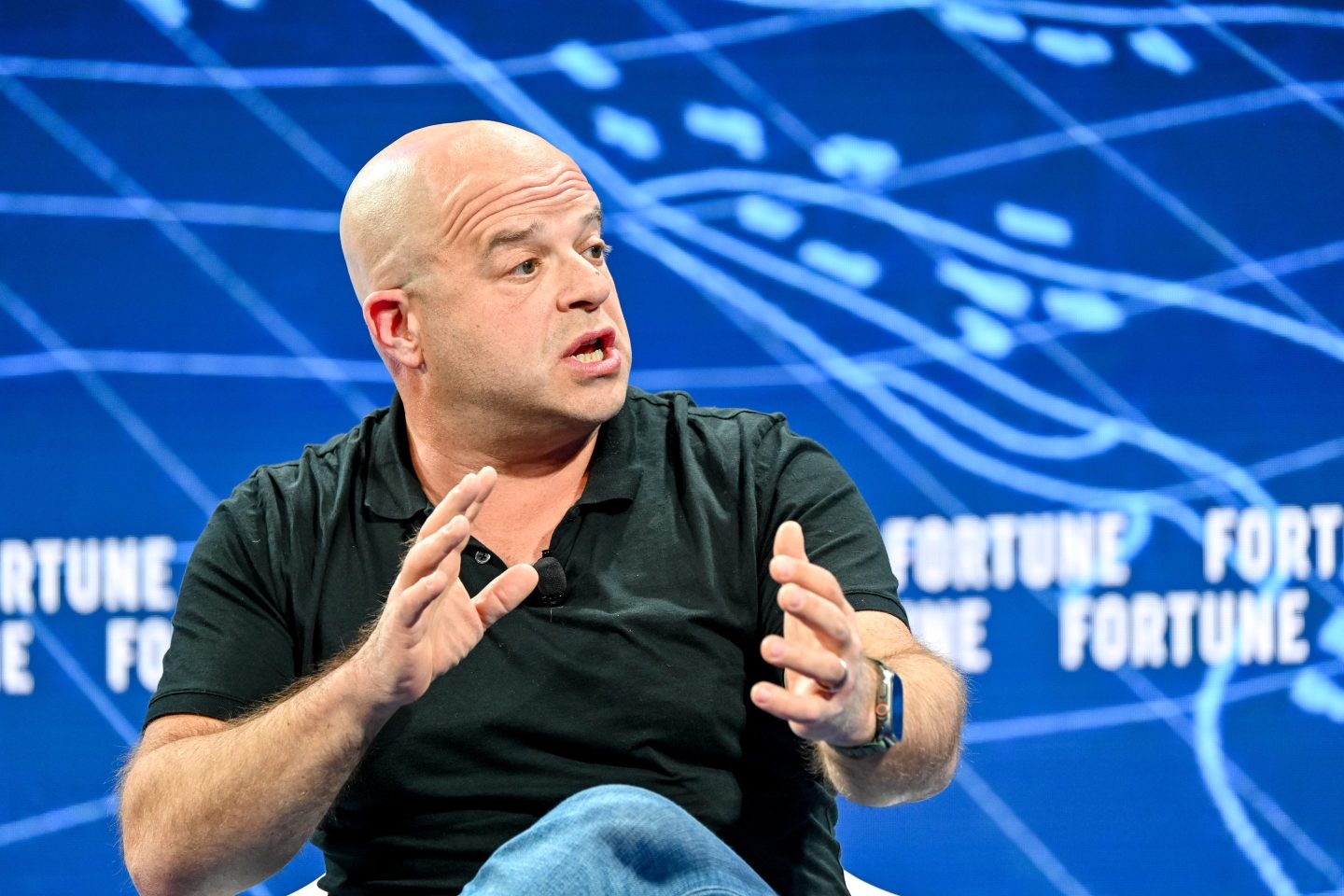 A man in a black polo and jeans gestures forward while sitting onstage.