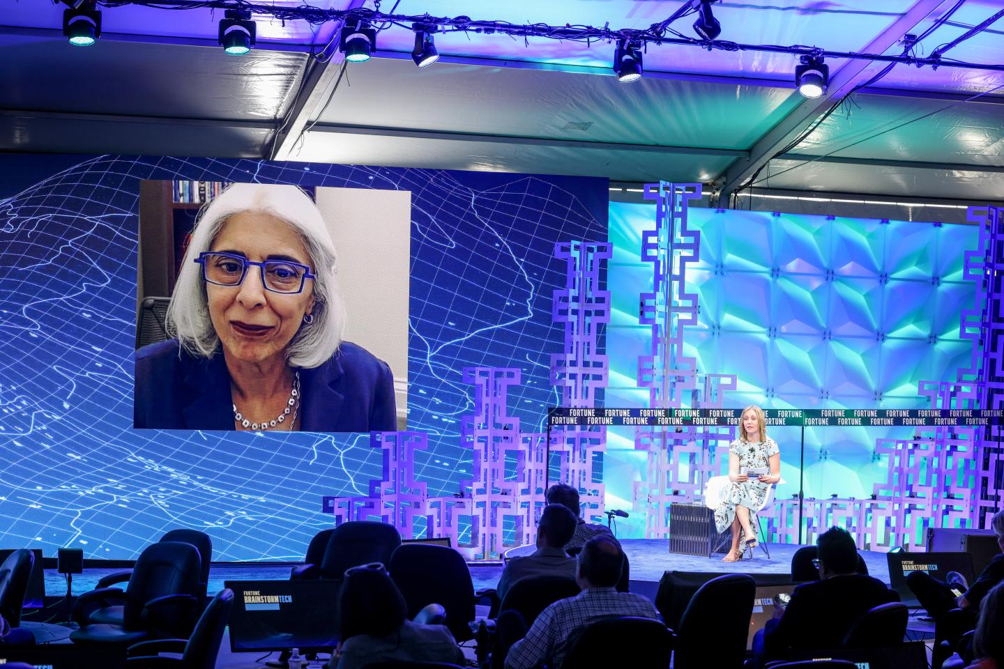 Dr. Arati Prabhakar is on the big screen on a conference stage, projected via Zoom. Below her is Fortune's EIC in a chair on stage, conducting the interview.