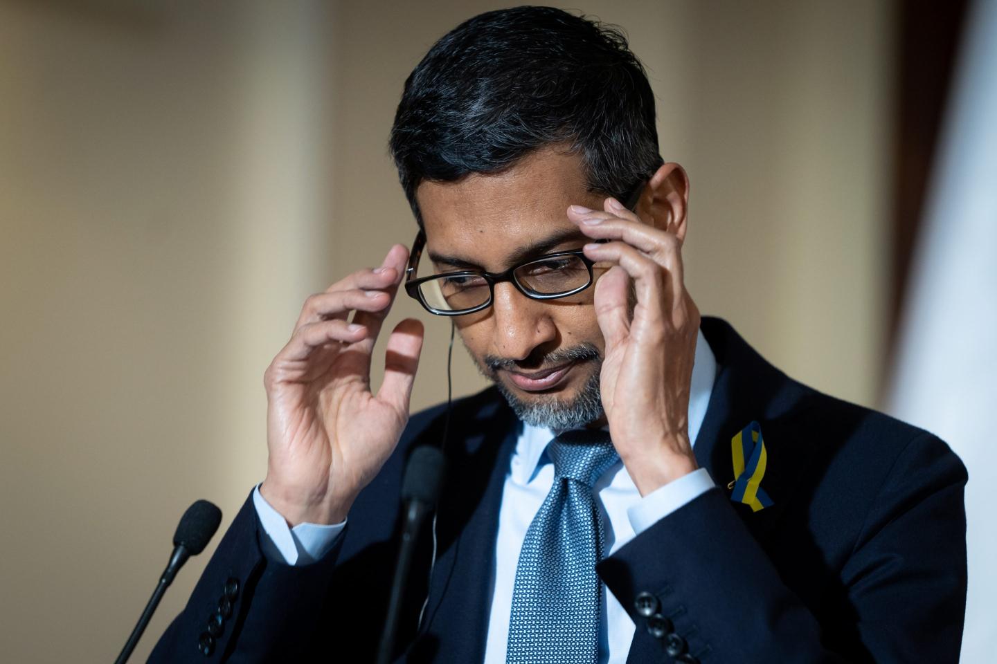 A man in a navy suit and blue tie adjusts his glasses at a podium with a microphone