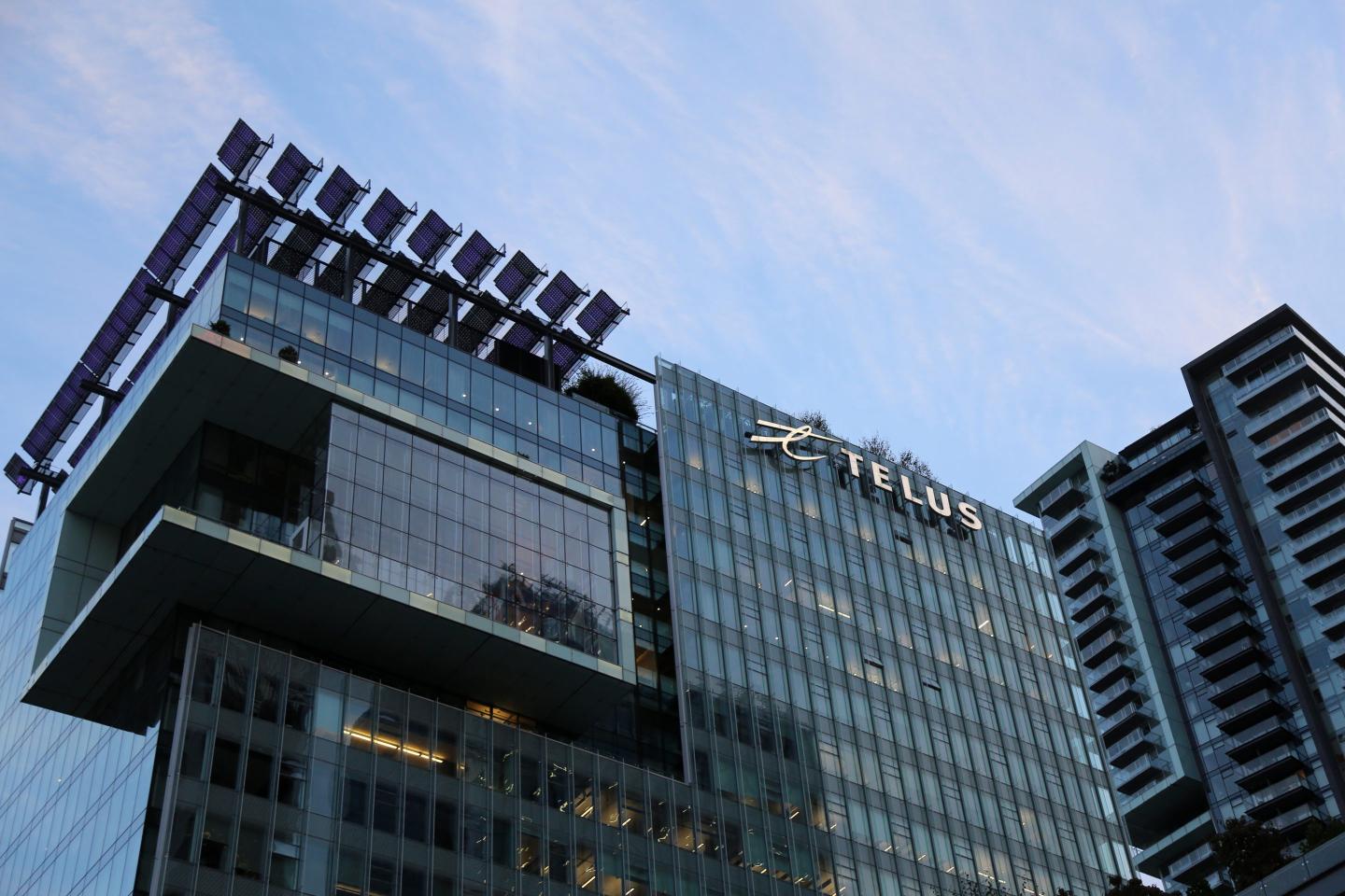 A glass and steel building towers against the blue sky. A sign reading "Telus" is mounted to the side of the building