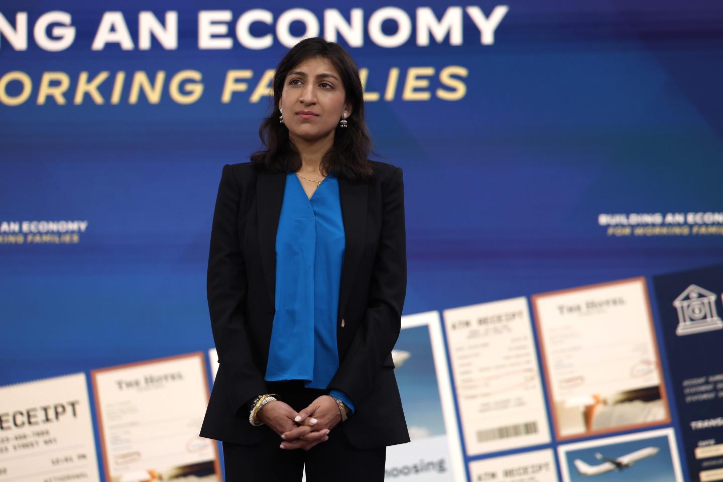 A woman in a blue blouse and back blazer stands upright with her hands folded in front of her on a stage