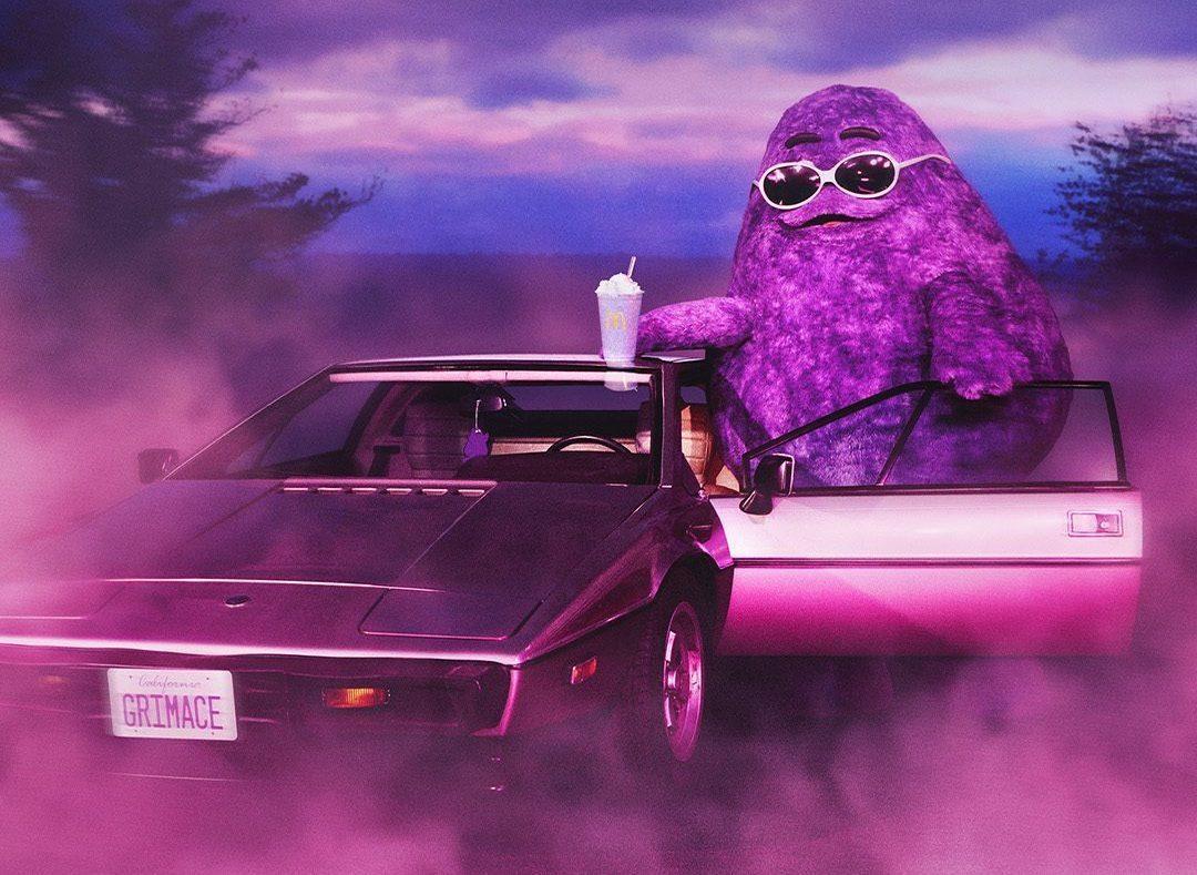 Grimace wearing sunglasses standing beside his car with a Grimace shake in hand.