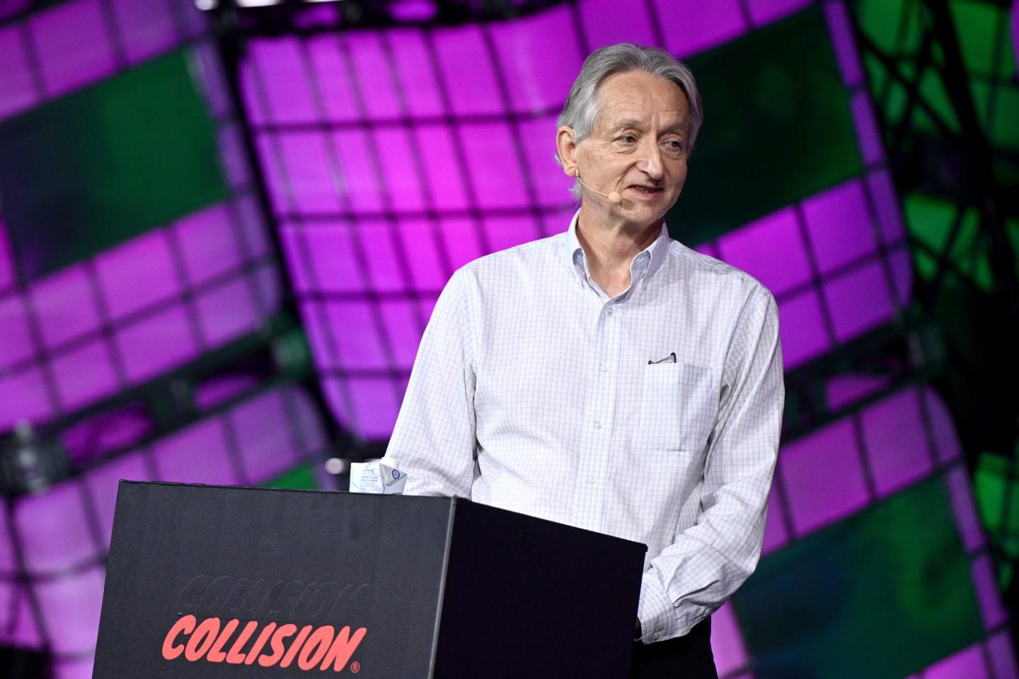 A man in a white collared shirt speaks from behind a podium onstage in front of a purple background