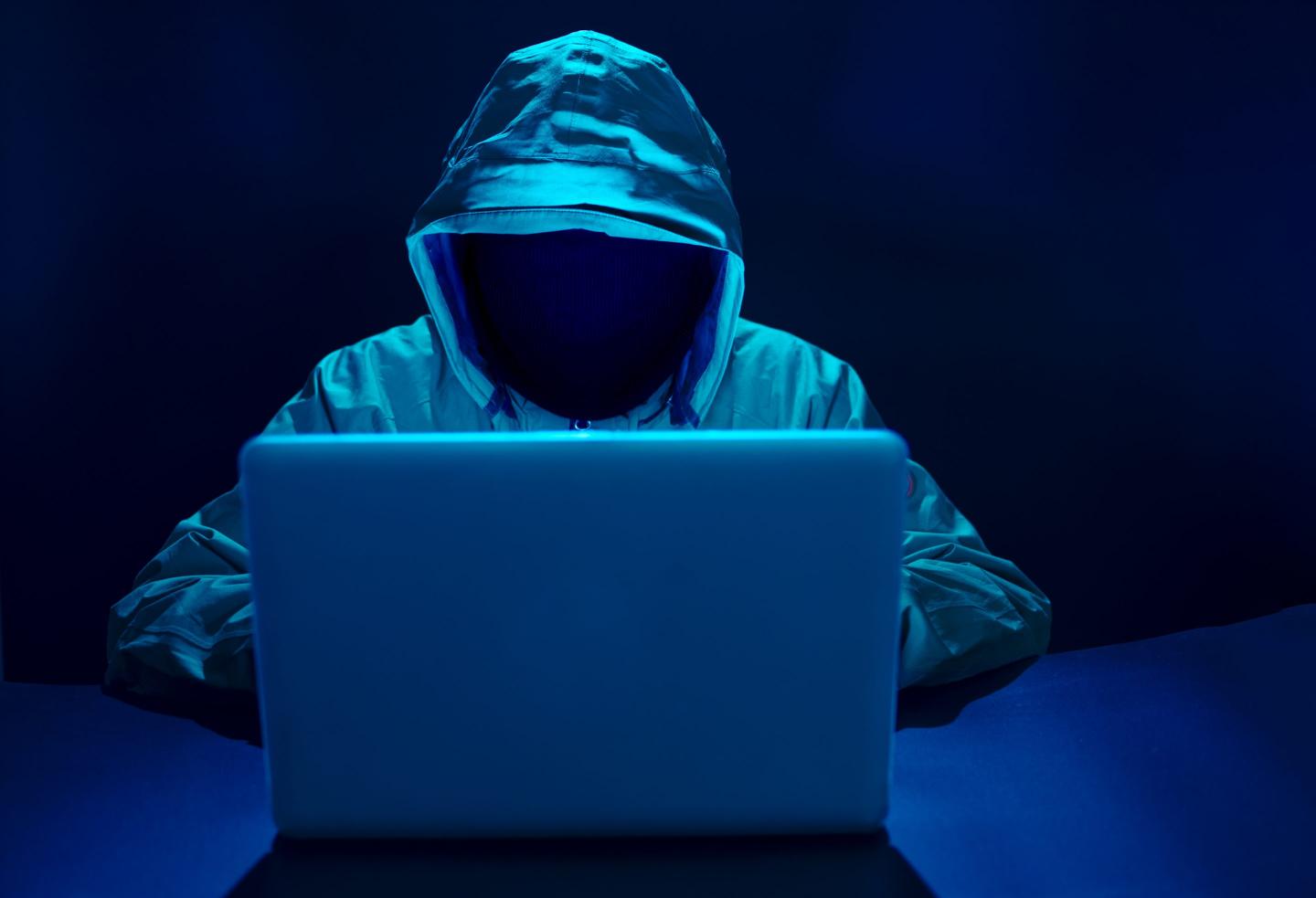 Hacker figure in hood sitting before an open laptop.