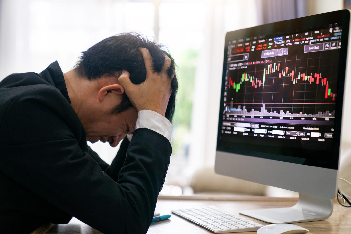 Stressed businessman, head in hands, sits before computer screen displaying stock market investments.