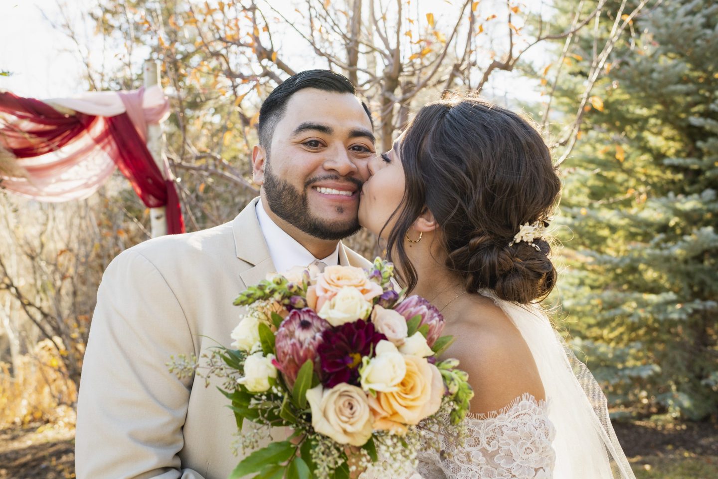 A bride kisses a groom on the cheek in a woodland setting.