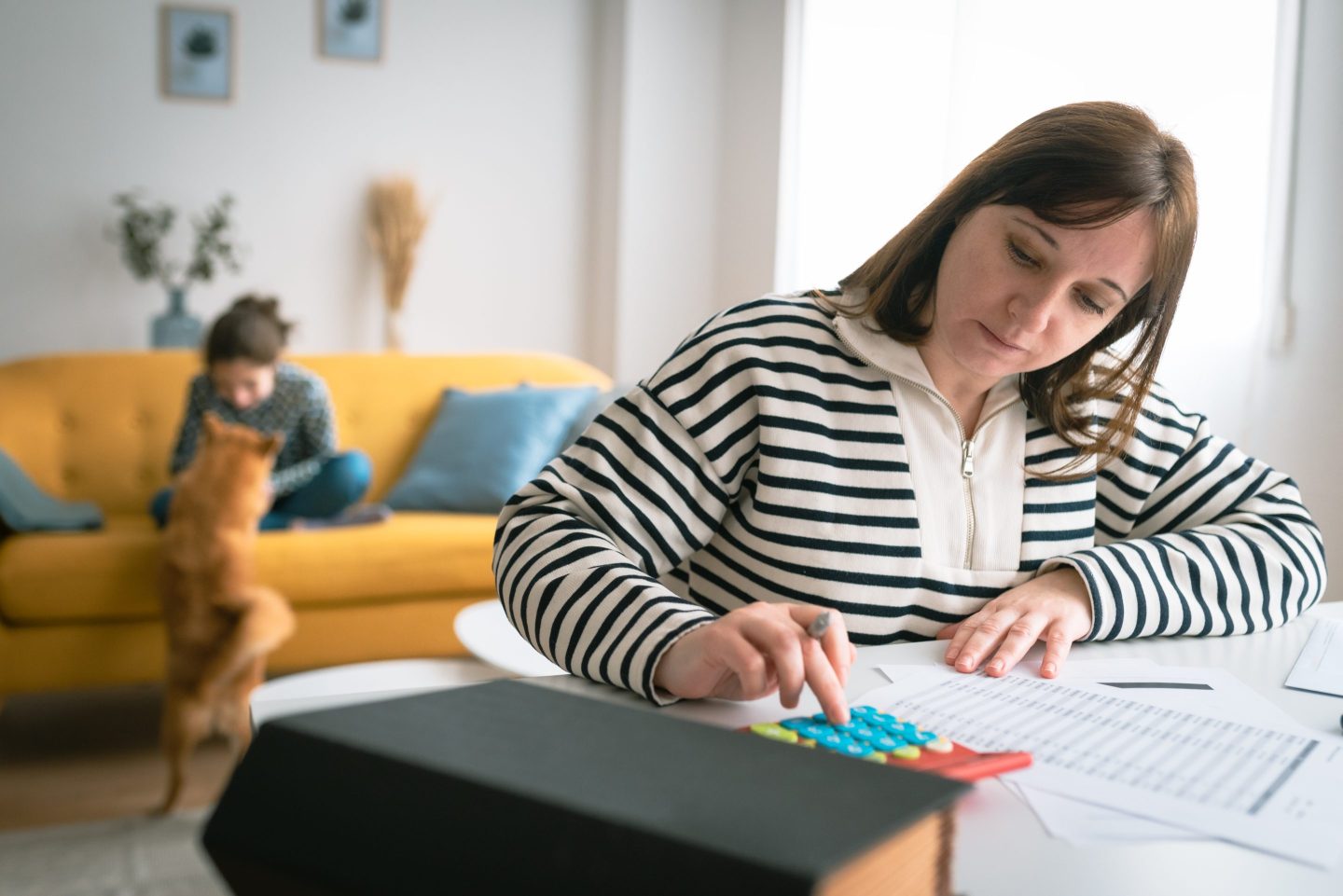 Mother manages family budget on calculator while her daughter plays with a dog in the background.